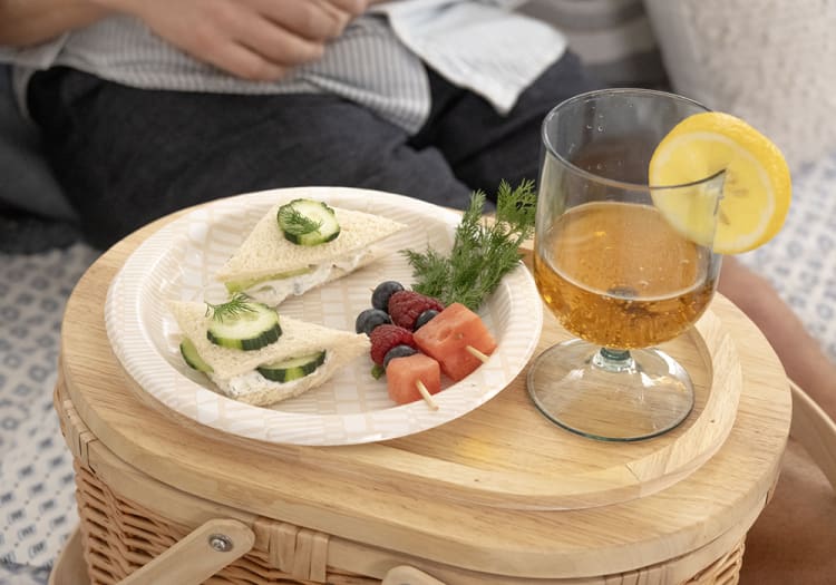 Small cucumber tea sandwiches and fresh fruit skewers on a patterned paper plate next to a glass of iced tea with a lemon slice on a wooden picnic basket.