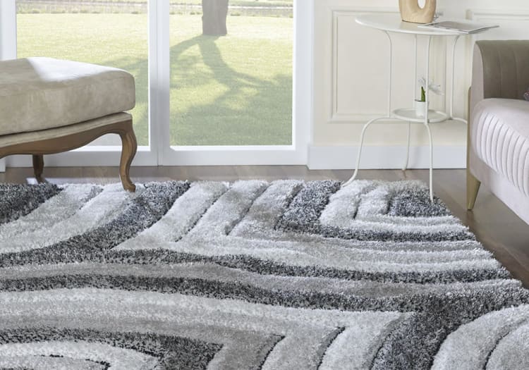 Gray and white geometric patterned shag area rug on hardwood floor, styled in a modern living room with beige upholstered furniture.
