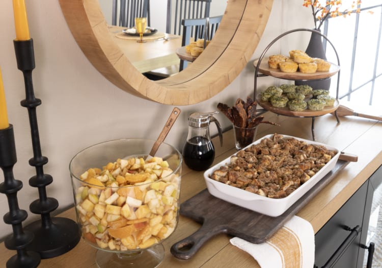 A wooden sideboard with a large round mirror above, featuring a glass bowl of mixed fruit salad, a white ceramic casserole dish with baked stuffing, a glass syrup dispenser, and a two-tiered stand holding savory muffins and bacon strips.