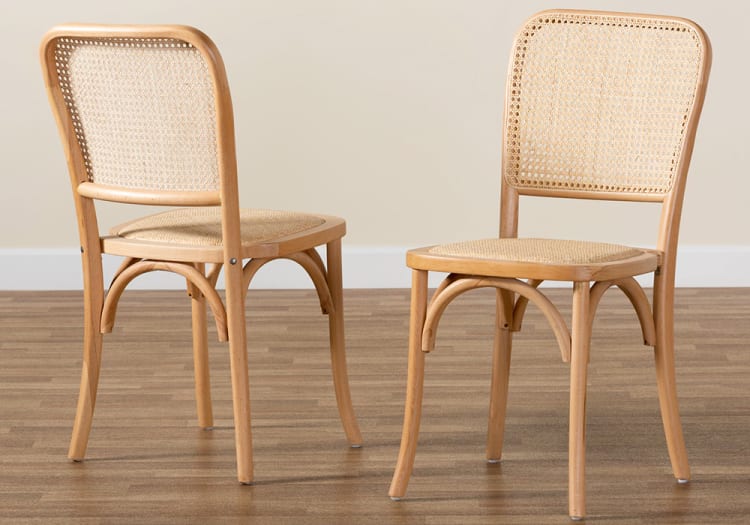 Two natural wood dining chairs with woven cane seats and backs, featuring curved legs and a light finish on a wooden floor.