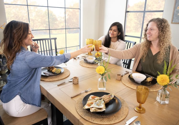 Three women sitting around a wooden dining table with woven placemats, clinking amber-colored glass goblets during a meal in a bright room with large windows.