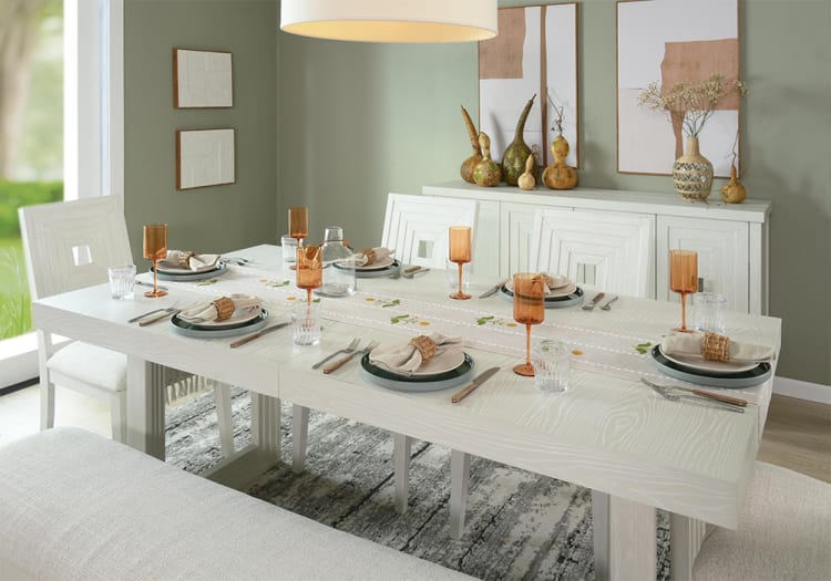 Modern dining room setup with a white wooden table, beige cushioned chairs, amber glassware, neutral napkins, and decorative gourds on a sideboard.