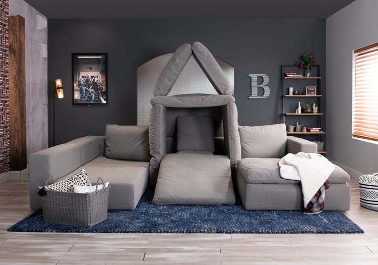 Modern living room with a gray sectional sofa arranged with cushions forming a house shape, set on a blue rug, featuring dark walls, a floor lamp, and a wooden shelf with decor.
