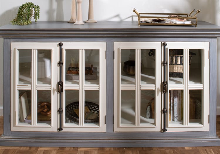 Gray wooden cabinet with cream-framed glass doors, featuring black metal handles, decorative items inside, and a plant and tray on top.