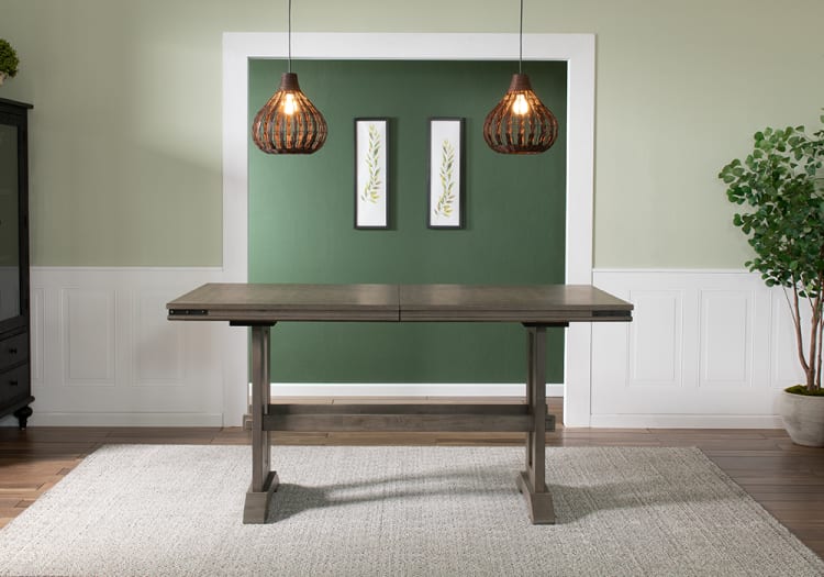Rustic gray wooden dining table with trestle legs, set against a green accent wall with two hanging wicker pendant lights and botanical wall art.