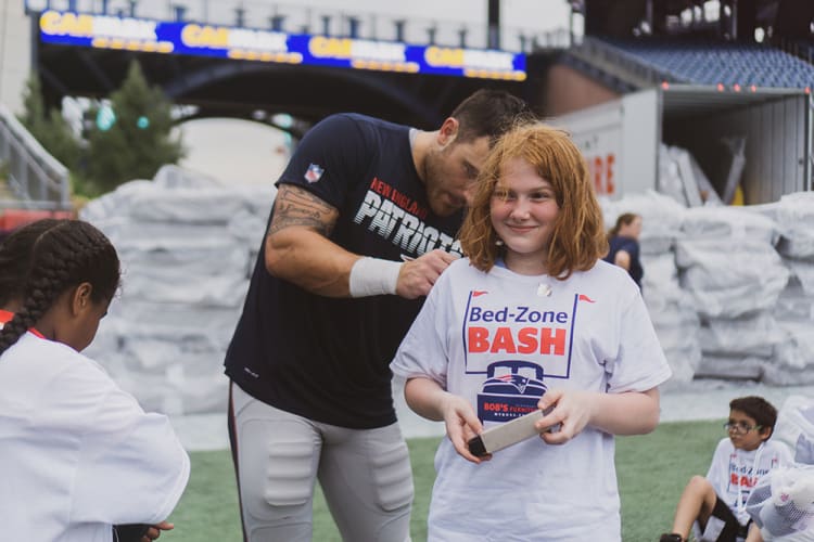 A person in a white "Bed-Zone BASH" t-shirt holding a box, with a man in a New England Patriots shirt signing an autograph on a sports field.