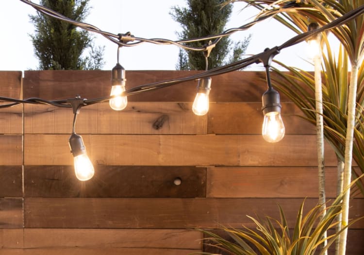 Outdoor string lights with warm glowing bulbs hanging against a wooden fence, creating cozy ambient lighting for patios or gardens.