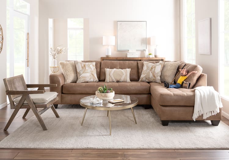 Modern living room with a tan leather sectional sofa, beige and patterned throw pillows, wooden armchair, round glass coffee table, and neutral decor.