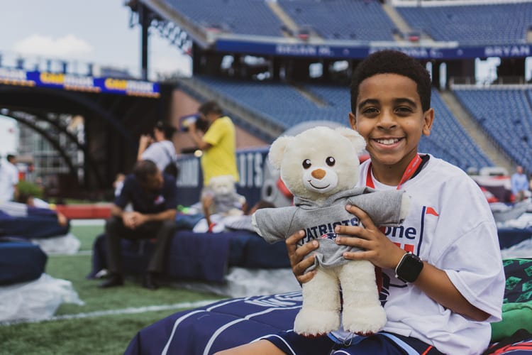 Young person sitting on a stadium field holding a white teddy bear wearing a gray Patriots hoodie, dressed in casual sportswear with a smartwatch.