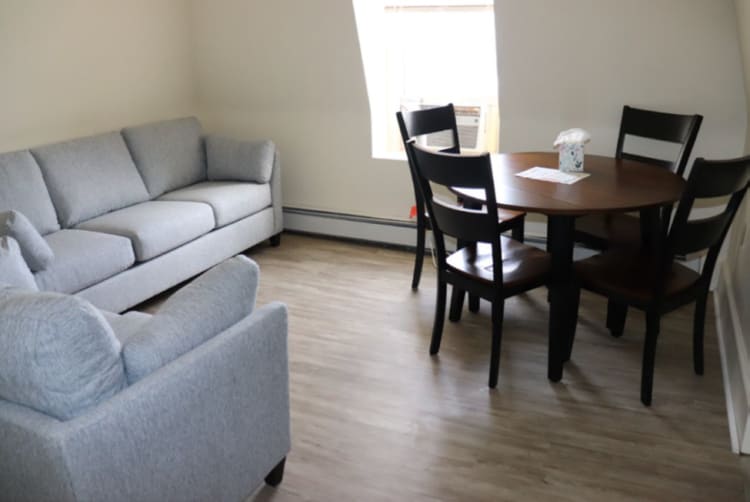 Modern living room with light gray fabric sofa and armchair, wood laminate flooring, and a round dark wood dining table with four black chairs.