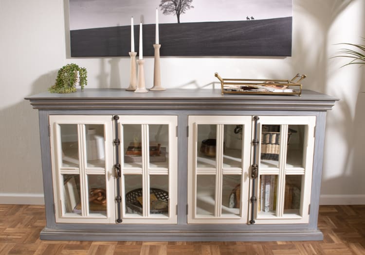 Gray and white wooden sideboard with glass paneled doors, decorative handles, and a top surface holding candles, a plant, and a gold tray.