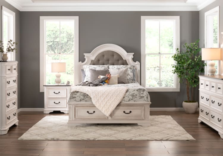 Bedroom with a white wooden bed featuring a tufted gray headboard, floral bedding, matching nightstands, dressers, beige rug, and natural light from windows.