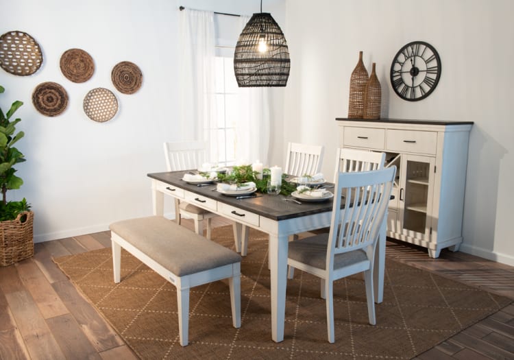 Dining room setup with white wooden table and chairs, gray cushioned bench, black tabletop, wicker pendant light, decorative woven wall baskets, and beige rug.