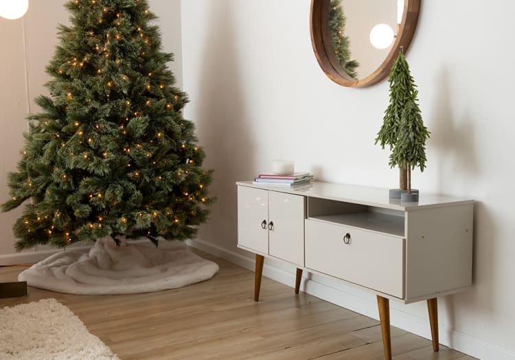 Decorated artificial Christmas tree with warm white lights next to a modern white mid-century style console table with wooden legs, small decorative tree, and stacked books.