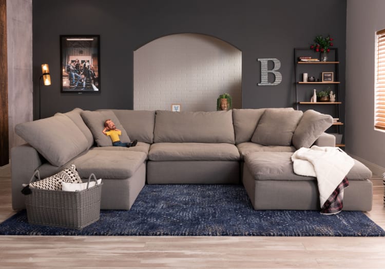 Spacious gray sectional sofa with plush cushions and adjustable backrest, placed on a blue patterned rug in a modern living room with dark accent wall and wooden shelves.