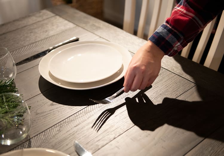 Hand placing a fork on a dark wooden dining table set with white ceramic plates, clear glassware, and silver cutlery, highlighting table setting for a meal.