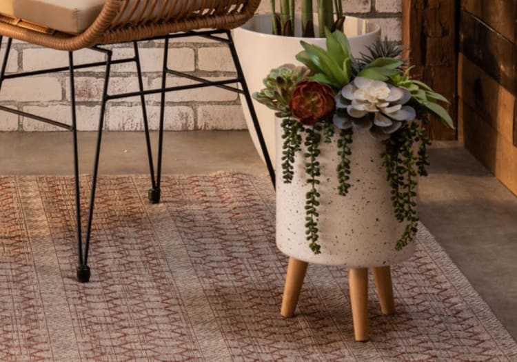 Modern speckled white ceramic planter with wooden legs, featuring assorted green and burgundy artificial succulents, placed on a patterned rug next to a wicker chair.