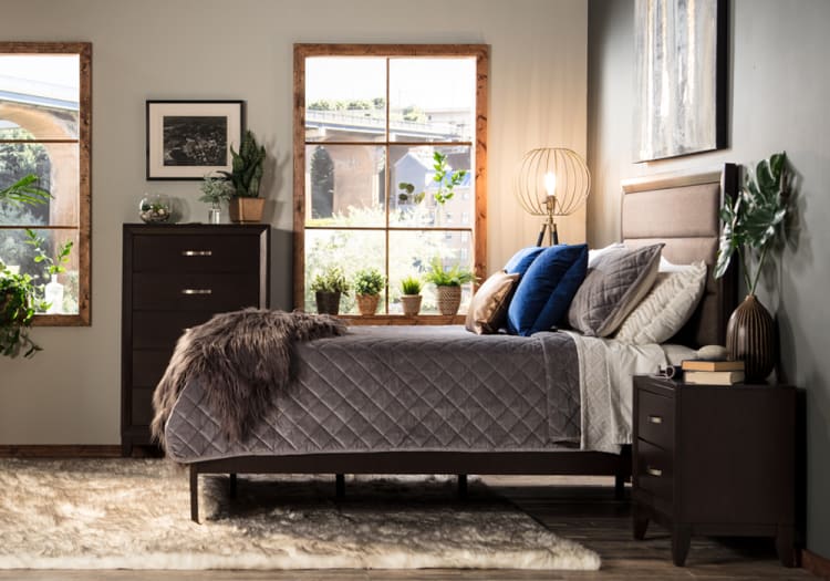 Modern bedroom with a dark wood bed frame, gray quilted bedding, blue and beige accent pillows, matching nightstand and dresser, and decorative plants.