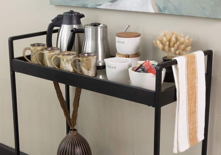 Black metal serving cart with four ceramic mugs, stainless steel coffee pots, white ceramic containers labeled "SUGAR" and "CREAM," a bowl with tea packets, and a white towel with an orange stripe.