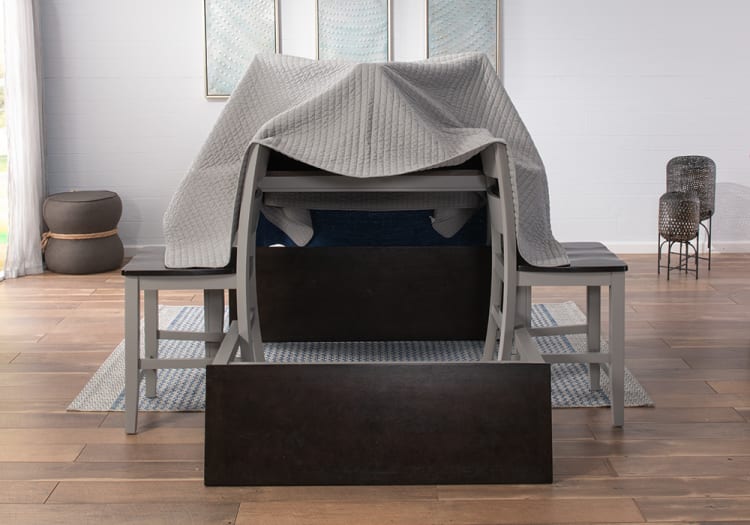 Gray and dark brown wooden dining table set with benches, covered by a gray quilted blanket, on a patterned rug in a modern room.