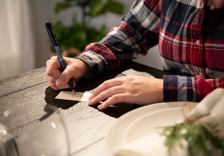 Person in a red and blue plaid shirt writing the word "You" on a small beige card with a black pen at a dark wooden table.