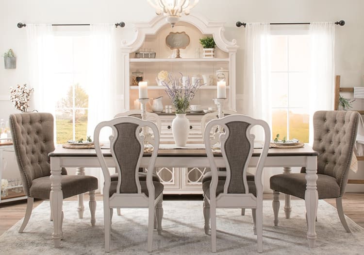 Dining room with a rustic white wooden table and six upholstered chairs in beige and gray fabric, featuring a white vase with lavender and lit candles.