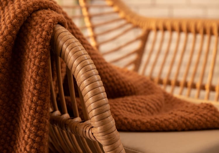 Brown knitted throw blanket draped over a natural rattan armchair with beige cushion, highlighting cozy home decor and textured fabric.