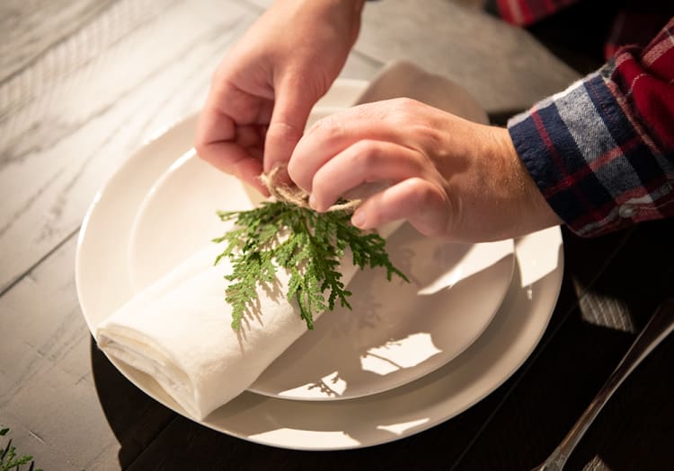 Hands tying a rustic twine around a white cloth napkin with green fern leaves on stacked white ceramic plates, set on a wooden table.