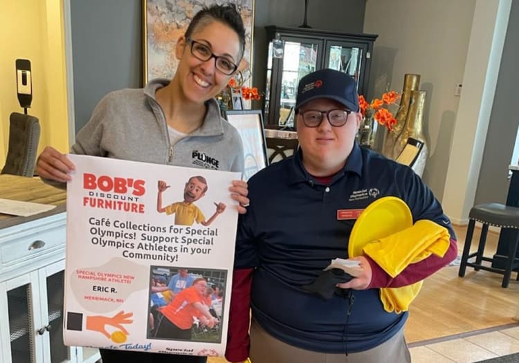 Two people holding a promotional poster for Bob's Discount Furniture supporting Special Olympics athletes. The poster reads: "BOB'S DISCOUNT FURNITURE Café Collections for Special Olympics! Support Special Olympics Athletes in your Community! SPECIAL OLYMPICS NEW HAMPSHIRE ATHLETE ERIC R. MERRIMACK, NH." One person wears a gray zip-up sweatshirt, and the other wears a navy blue polo with a Special Olympics logo, holding yellow cloth and plates. The setting is a home interior with furniture and decor.