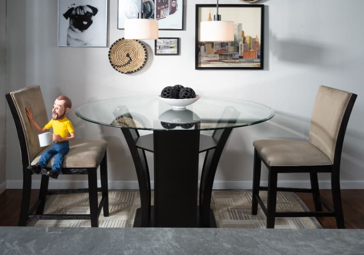Modern dining area with a round glass-top table supported by a black base, two beige cushioned chairs, pendant lights, and wall art including a dog portrait.