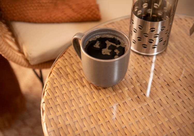 Gray ceramic coffee mug filled with black coffee on a round wicker table next to a stainless steel French press with coffee bean cutouts.