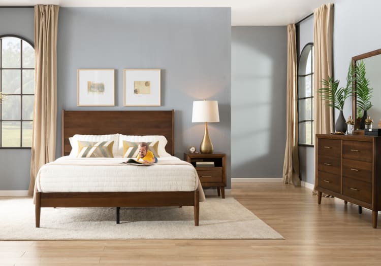 Modern bedroom with gray walls, wooden bed frame and matching nightstand, beige curtains, large windows, and wooden dresser with mirror and green plants.