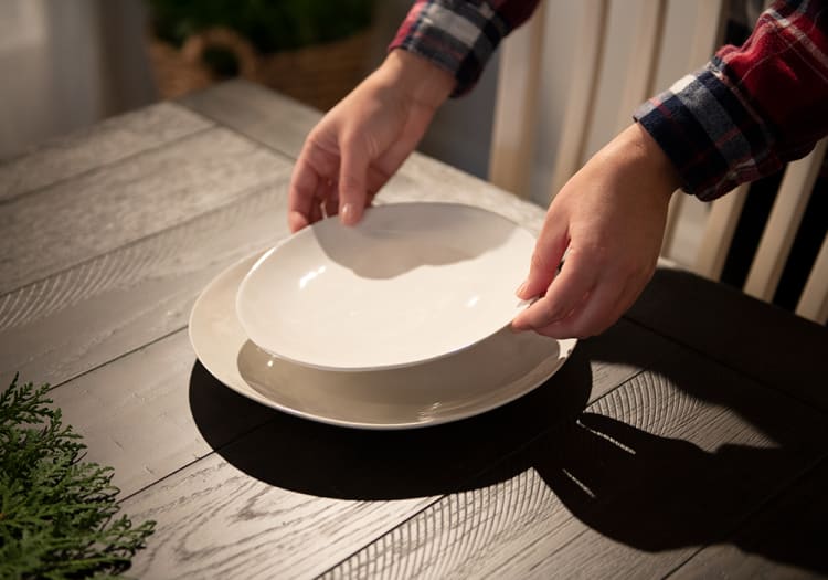 Hands placing a white ceramic plate on top of another on a dark wooden dining table, highlighting table setting and dinnerware.