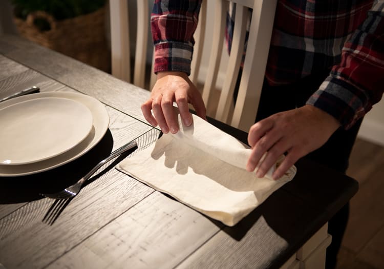 Hands folding a beige cloth napkin on a dark wooden dining table next to stacked white ceramic plates and silverware.