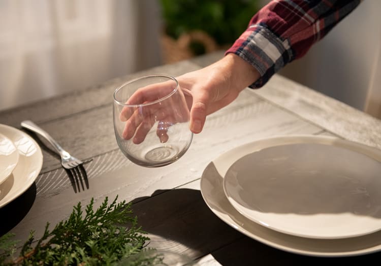 Hand holding a clear, round glass tumbler above a rustic wooden dining table set with white ceramic plates and a silver fork, with green foliage as table decor.