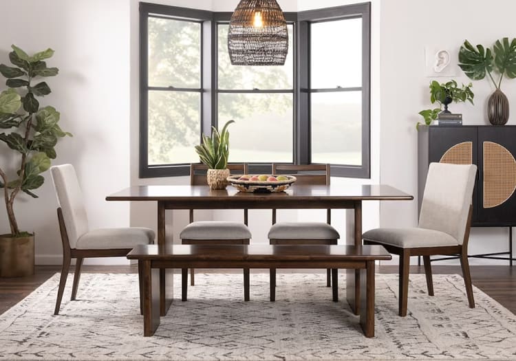 Modern dining room set with dark wood rectangular table, matching bench, and beige upholstered chairs on patterned rug, near bay window.