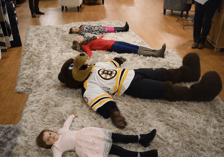 Three children and a person in a Boston Bruins bear mascot costume lie on a large beige shag rug on a wooden floor, showcasing playful indoor activity and team spirit.