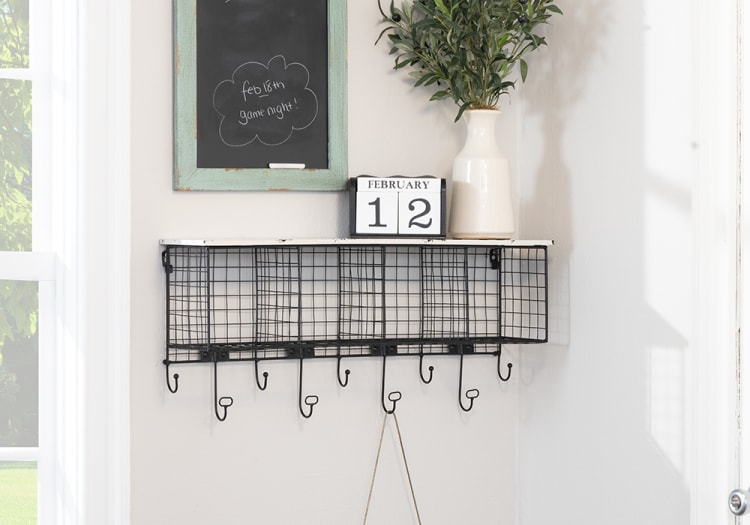 Wall-mounted black metal wire organizer with six compartments and hooks, topped with a white ceramic vase with greenery and a wooden calendar showing February 12. A small chalkboard above reads "feb 18th game night!"