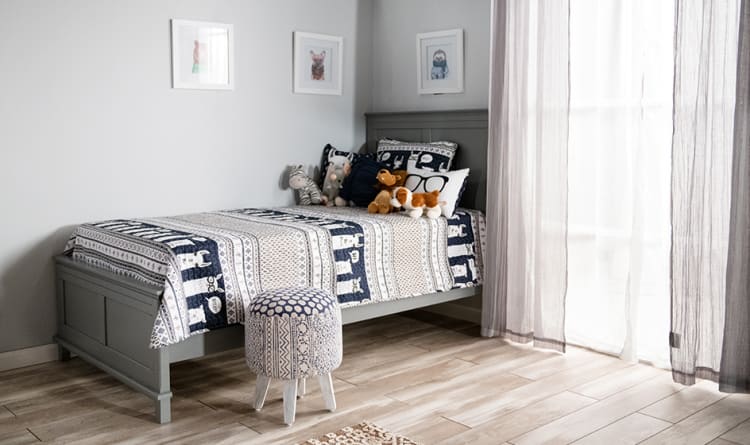 Single gray wooden bed with patterned navy and beige bedding, assorted plush toys, decorative pillows, and a blue patterned stool in a bright, minimalist bedroom.