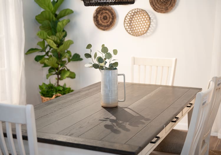 Dining room with a dark wood rectangular table, white wooden chairs, a ceramic pitcher vase with eucalyptus, and a green potted plant.