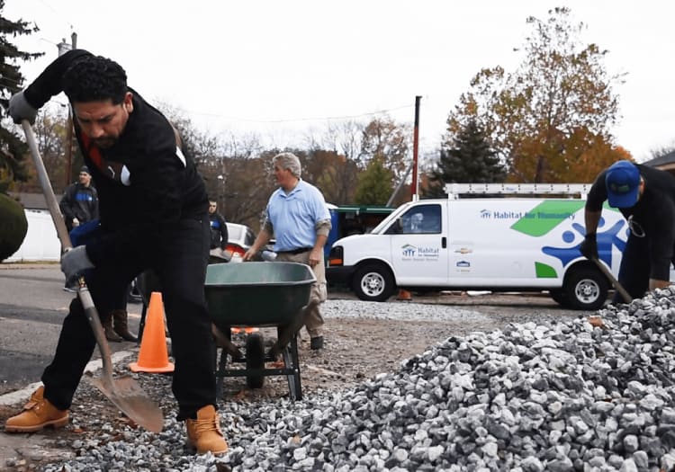 Volunteers working with shovels and wheelbarrow to move gravel for Habitat for Humanity construction project, wearing work gloves and boots.