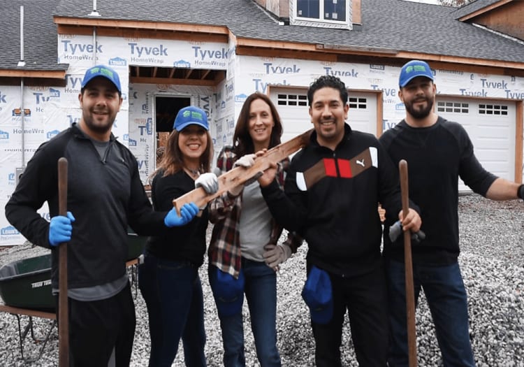Group of five construction workers wearing gloves and blue hats holding wooden planks and tools in front of a house under construction with Tyvek HomeWrap visible.