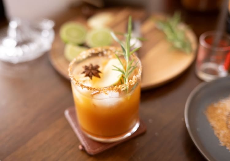Glass of orange-colored cocktail garnished with a rosemary sprig, placed on a brown leather coaster on a wooden table.