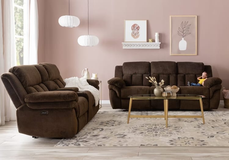 Two dark brown plush fabric sofas with tufted backs and armrests arranged in a living room with a beige patterned rug, glass coffee table with gold legs, and minimalist wall art on a soft pink wall.