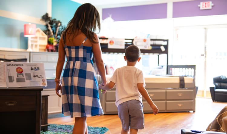 A woman in a blue and white checkered dress holding hands with a child in a white shirt and gray shorts, walking inside a furniture store.
