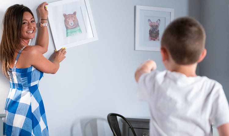 A woman in a blue and white checkered dress hangs a framed bear illustration on a gray wall while a child in a white shirt points toward the wall.