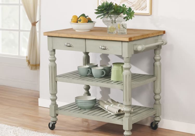 Kitchen cart with light gray painted wooden frame, natural wood top, two drawers, two slatted shelves, and caster wheels, styled with bowls, mugs, and a bowl of vegetables.