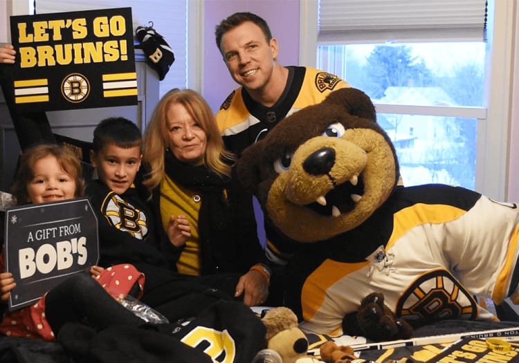 Group wearing Boston Bruins apparel holds signs reading "LET'S GO BRUINS!" and "A GIFT FROM BOB'S" alongside Bruins mascot in a room with large windows.