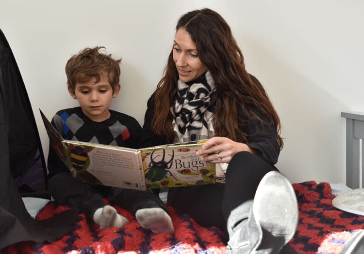 A woman and a child sitting on a bed with a red and black blanket, reading the book "Beetles and Bugs" together.