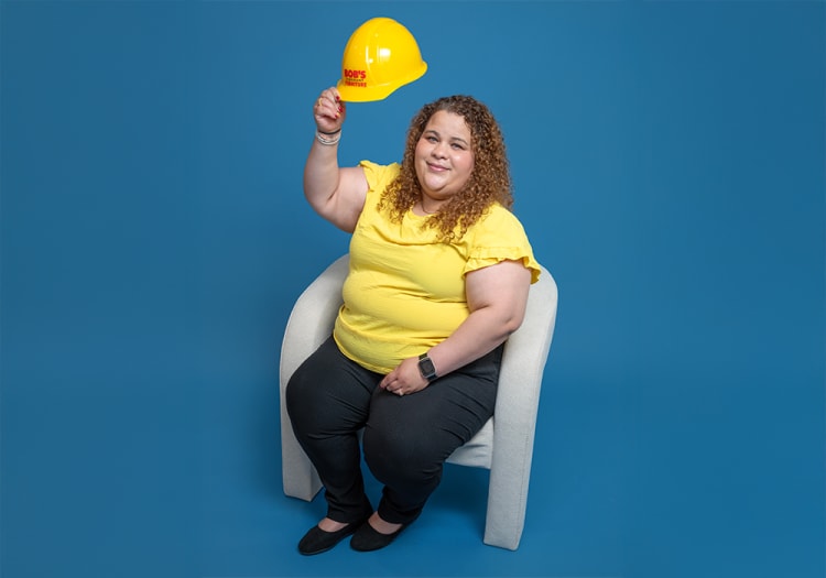 Person wearing a yellow ruffled short-sleeve top and black pants sitting on a white chair, holding a yellow hard hat with "BOB'S" text.